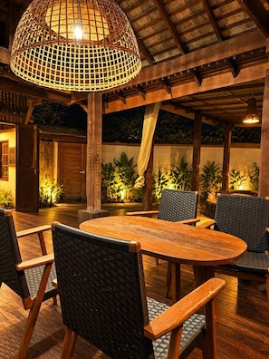 An outdoor dining area with a round wooden table and wicker chairs, under a wooden pergola with a woven light fixture. - Chaba Varee Resort Doi Saket Chiang Mai (Pa Meung)