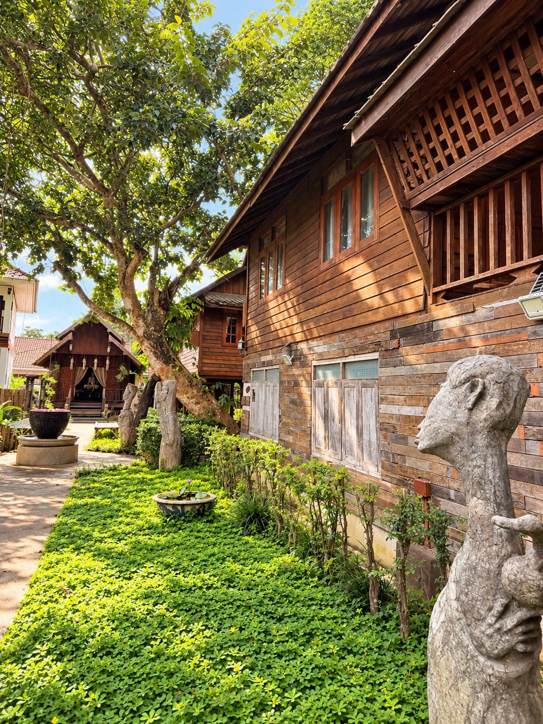A wooden house with a porch, surrounded by a well-maintained garden with various plants and a stone sculpture.