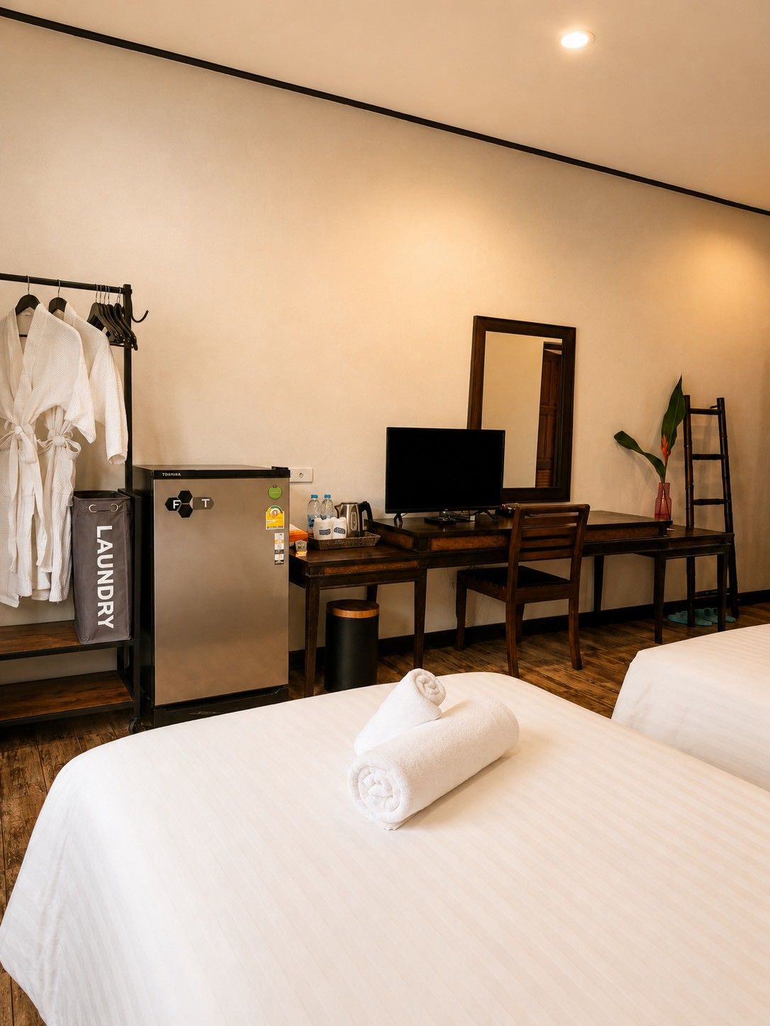 Ein Hotelzimmer mit zwei Betten, einem Schreibtisch mit Computer, einem kleinen Kühlschrank, einem Spiegel, einer Pflanze und einem Wäschewagen.