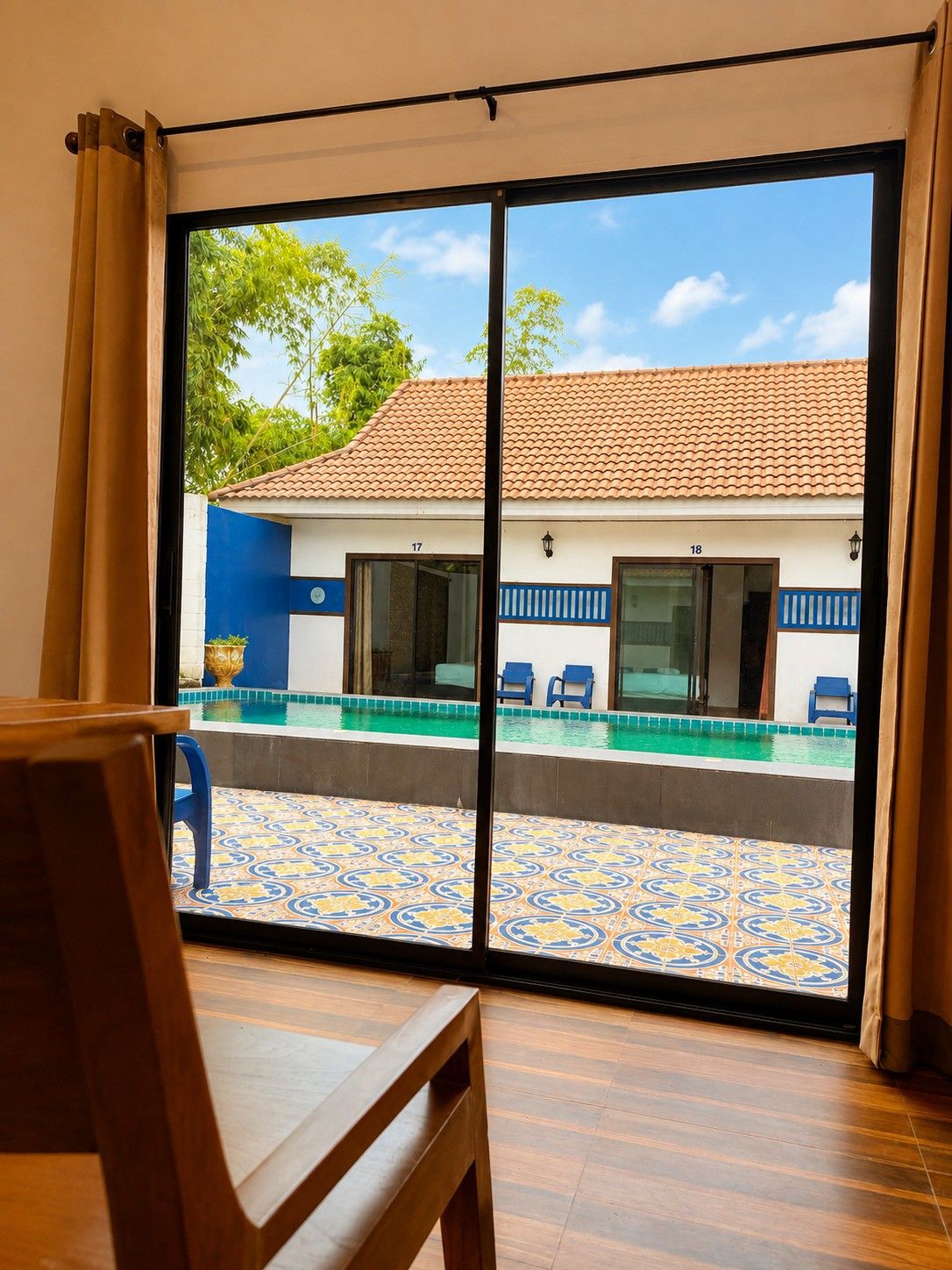 Quarto com uma grande porta de correr de vidro que dá para uma área de piscina com cadeiras azuis e piso de azulejos.