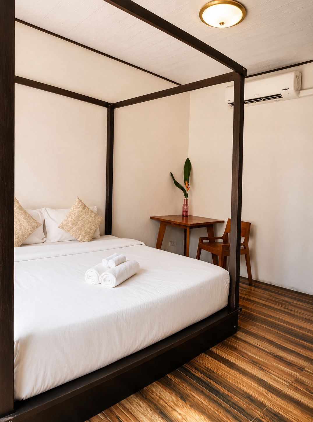 Una cama con dosel con sábanas blancas, una mesita con un jarrón y una silla, y un equipo de aire acondicionado en la pared.