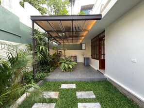 Un espacio exterior techado con una mesa y sillas, rodeado de follaje y un sendero de piedra.