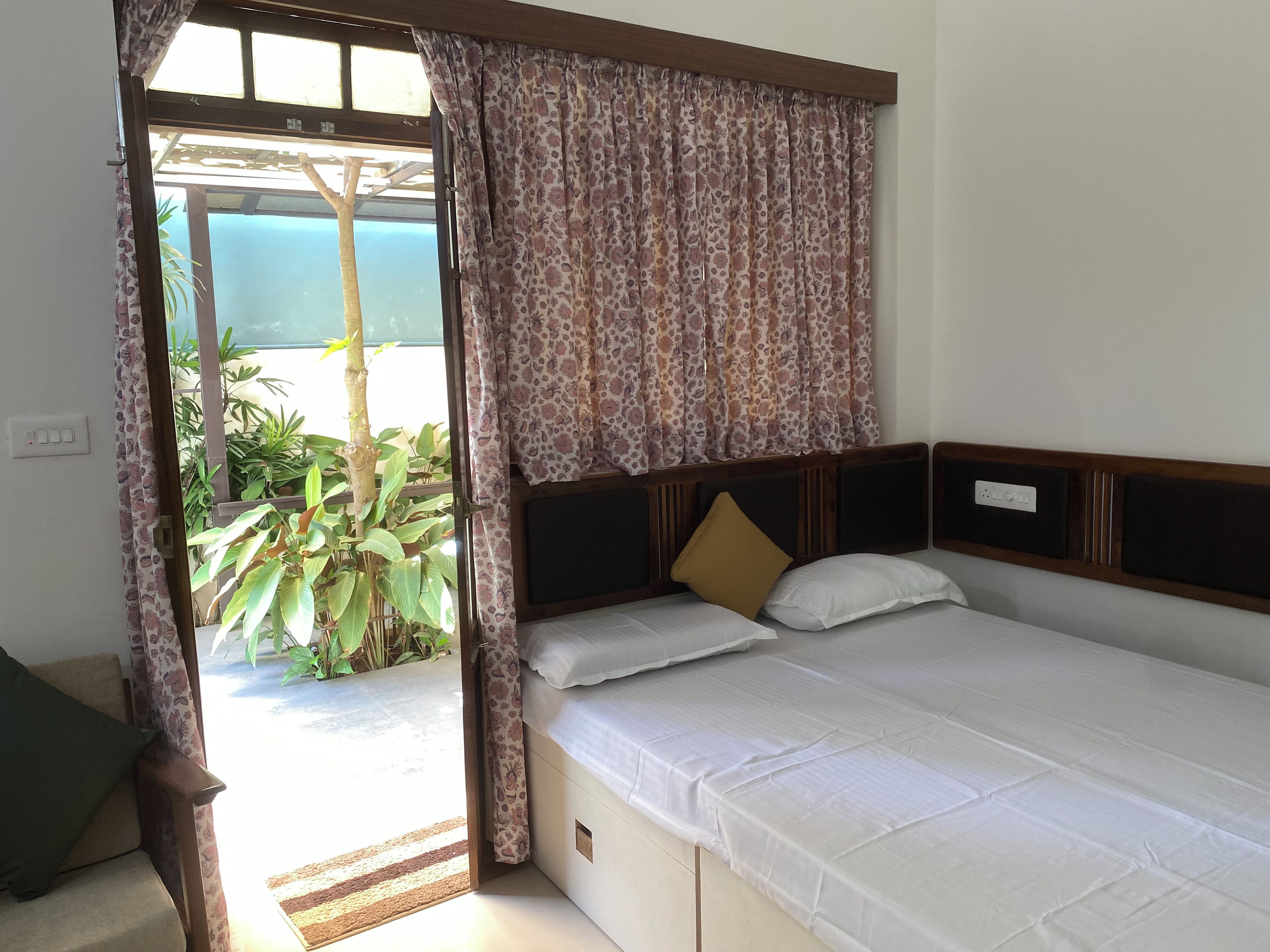 Una habitación con cama, ventana con cortinas y vistas al exterior con vegetación, además de un enchufe en la pared.