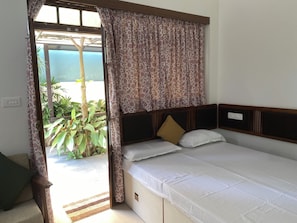 A room with a bed, a window with curtains, a view of greenery outside, and a power outlet on the wall.