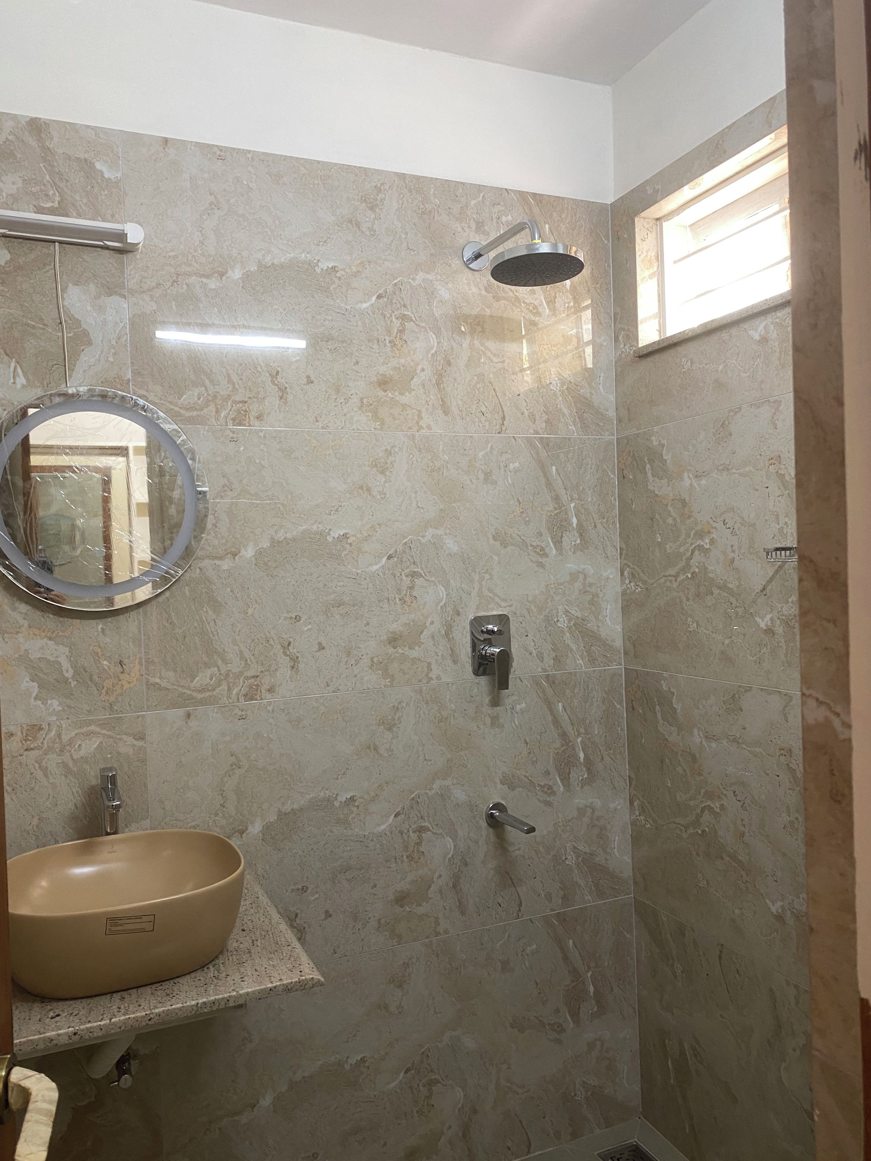 Une salle de bain avec un mur en marbre, un miroir rond, un lavabo beige, une douchette à main et une fenêtre.