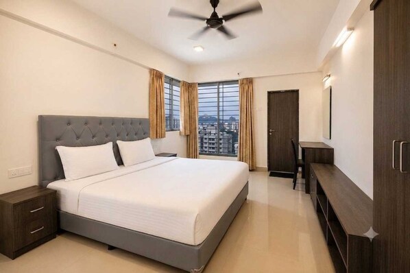 A hotel room with a bed, a desk, a chair, a wardrobe, and a ceiling fan. - Suite Hotel Urban Oasis (Pune)