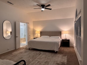 Un dormitorio con cama, dos mesitas de noche con lámparas, ventilador de techo, espejo grande y puerta que da a un balcón.