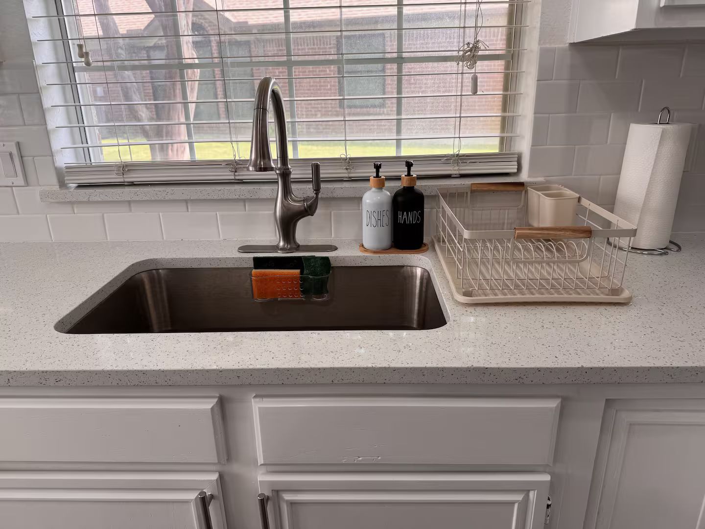 Una cocina con un lavamanos de acero inoxidable, una ventana con persianas y una encimera con un escurreplatos y dos botellas con la etiqueta 'DISHES' y 'HANDS'.
