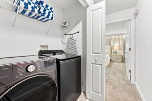 Ein Waschraum mit einer Frontlader-Waschmaschine und einem Trockner, ein Wäscheständer mit Handtüchern und eine Tür zu einem Schlafzimmer mit Bett und Nachttisch.