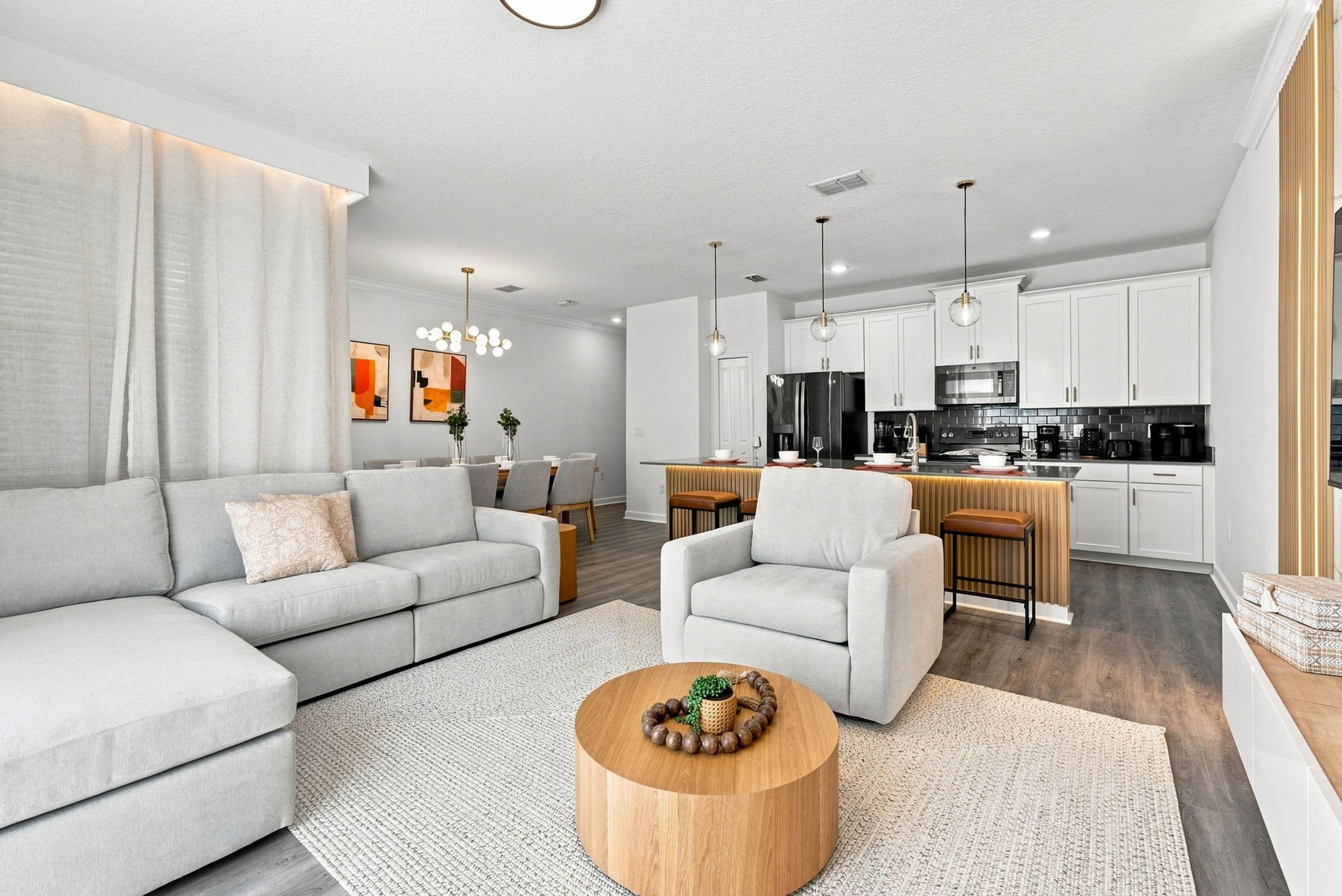 A modern living room with a sofa, armchair, and coffee table, a dining area with chairs, and a kitchen with appliances.