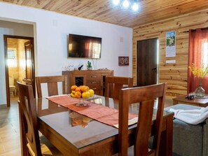Una zona de menjador amb taula de fusta, cadires, un plat de fruita, un televisor i una paret de fusta amb una porta.