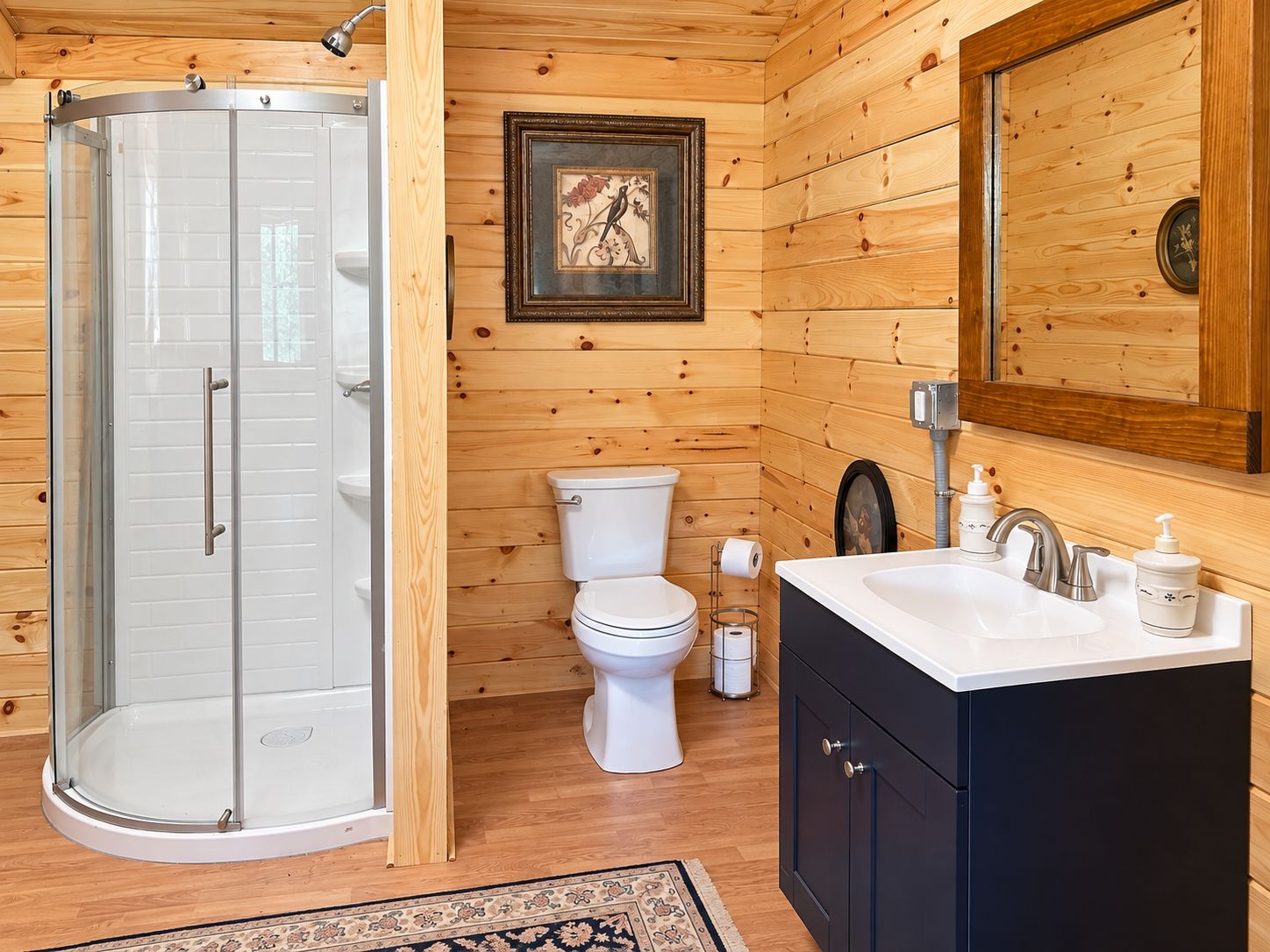 A wooden cabin bathroom with a glass shower enclosure, a white toilet, a sink with a mirror, and a framed picture on the wall.