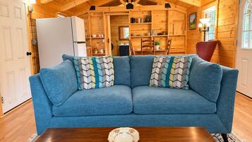 A cozy room with a blue sofa, wooden walls, and a dining area visible through an open door.