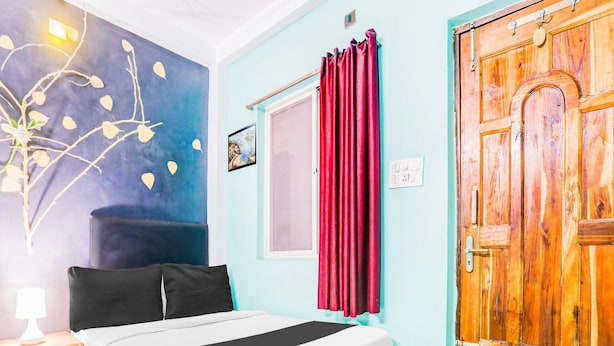 Une chambre à coucher avec un lit, une table de chevet, un mur orné d’un tableau, une fenêtre avec des rideaux et une porte en bois.