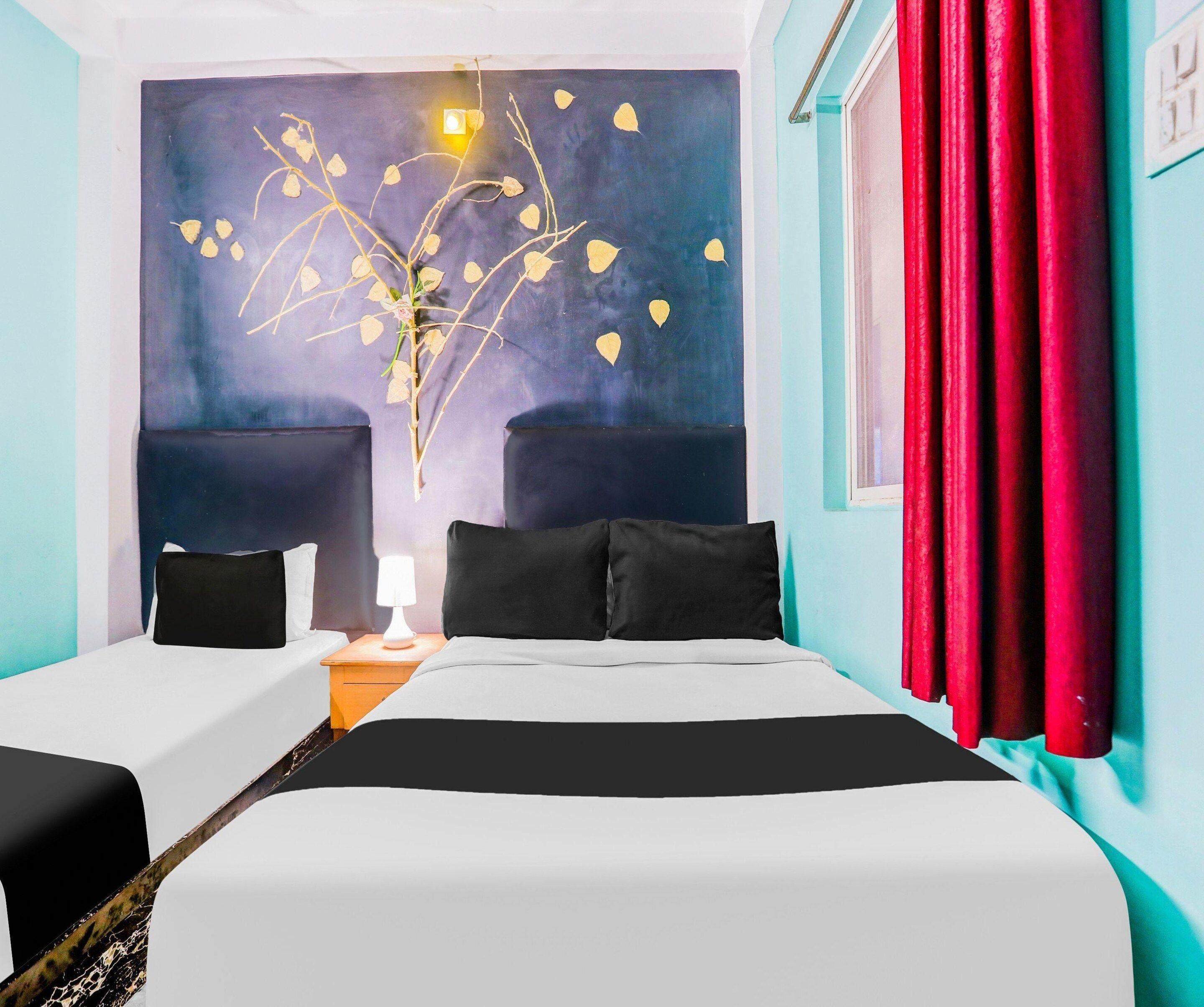 Een hotelkamer met twee bedden, een nachtkastje met een lamp, een muur met een decoratieve plant en een raam met rode gordijnen.