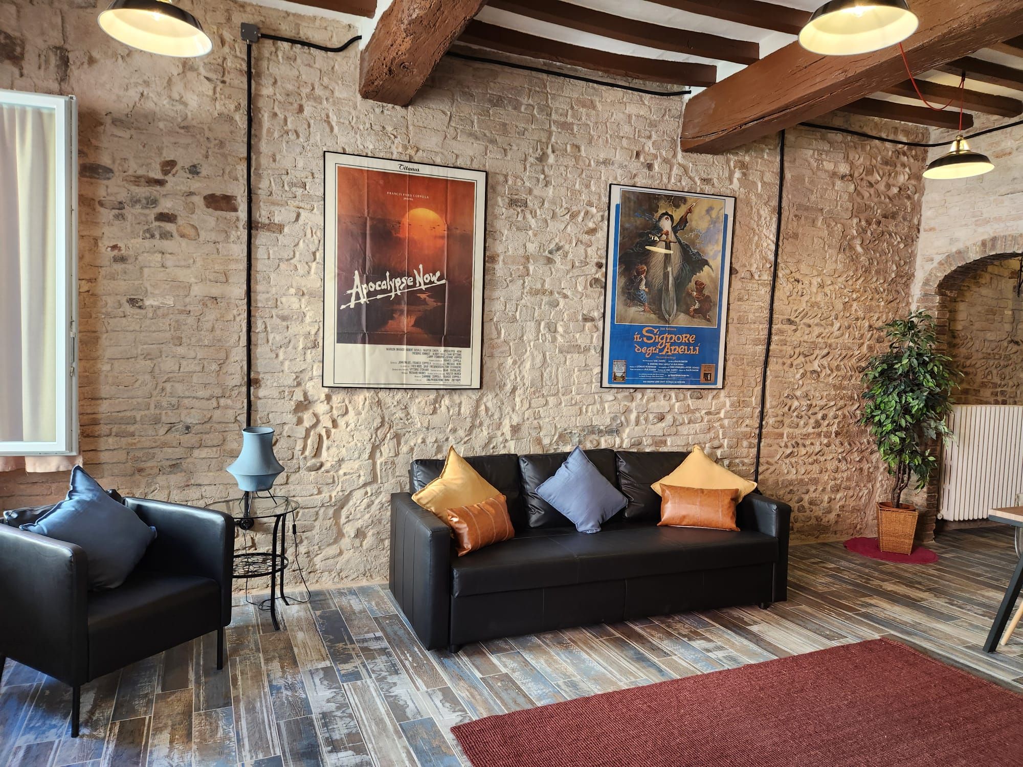 Una zona de estar acogedora con un sofá de cuero negro, dos sillones y una alfombra roja. Hay dos carteles en la pared de ladrillo, uno de los cuales se titula 'Apocalypse Now'.