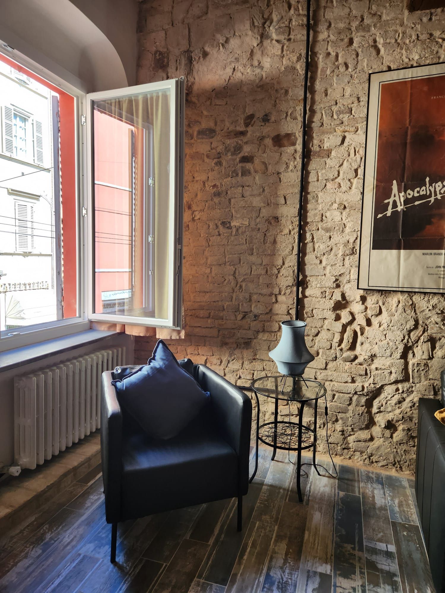 Ein Zimmer mit Backsteinwand, einem Fenster mit geöffneten Fensterläden, einem schwarzen Ledersofa, einem kleinen Tisch mit Lampe und einem Poster an der Wand.