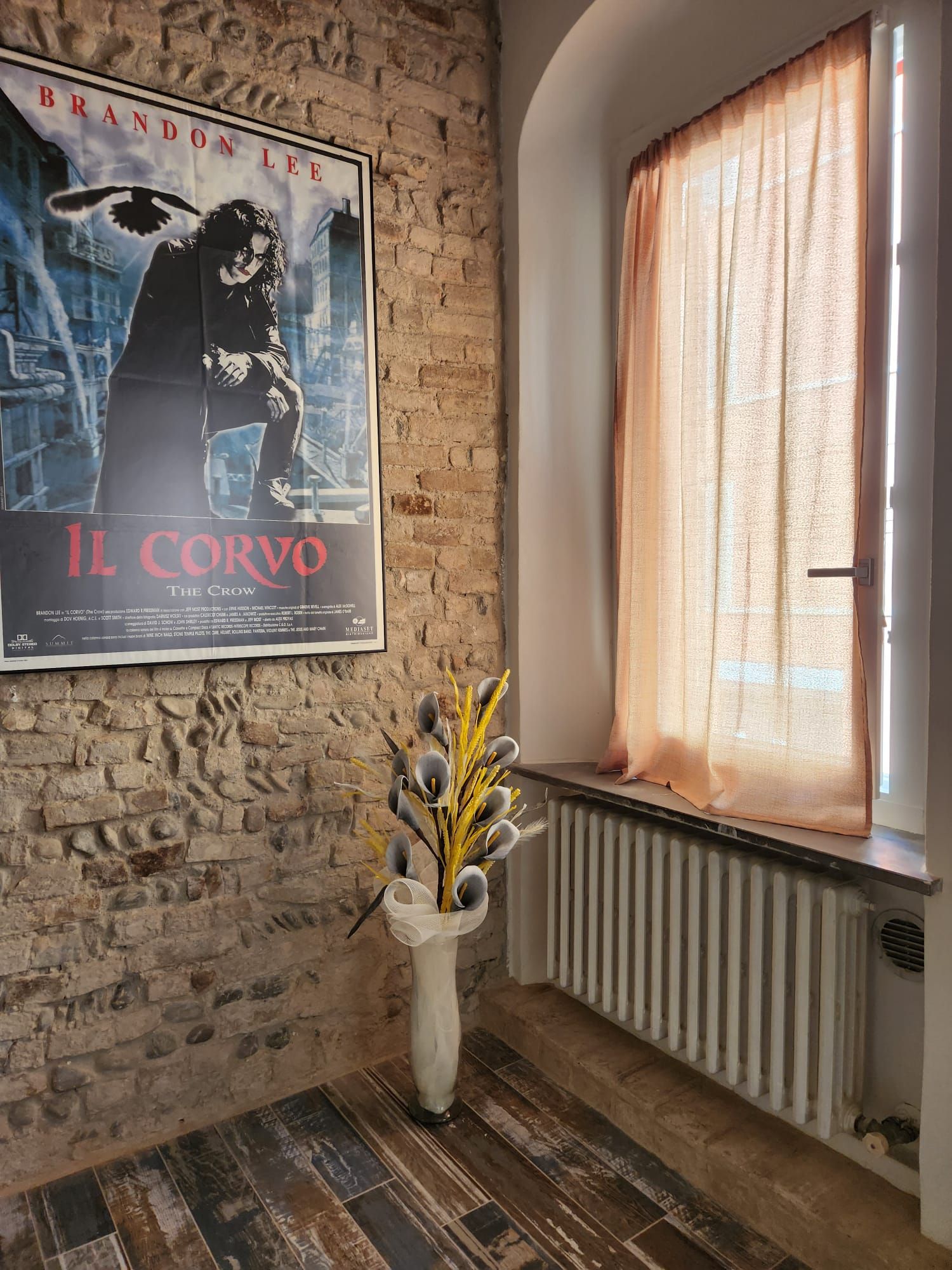 Ein Zimmer mit einem Poster an der Wand, einem Fenster mit Vorhängen und einer Vase mit Blumen auf dem Boden.