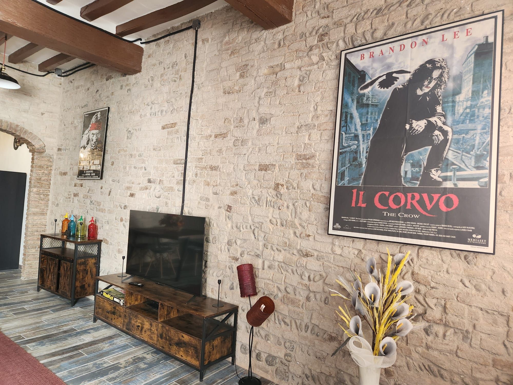 Una habitación con pared de ladrillo, un cartel de 'Il Corvo', un armario de madera, un televisor y un florero con flores.
