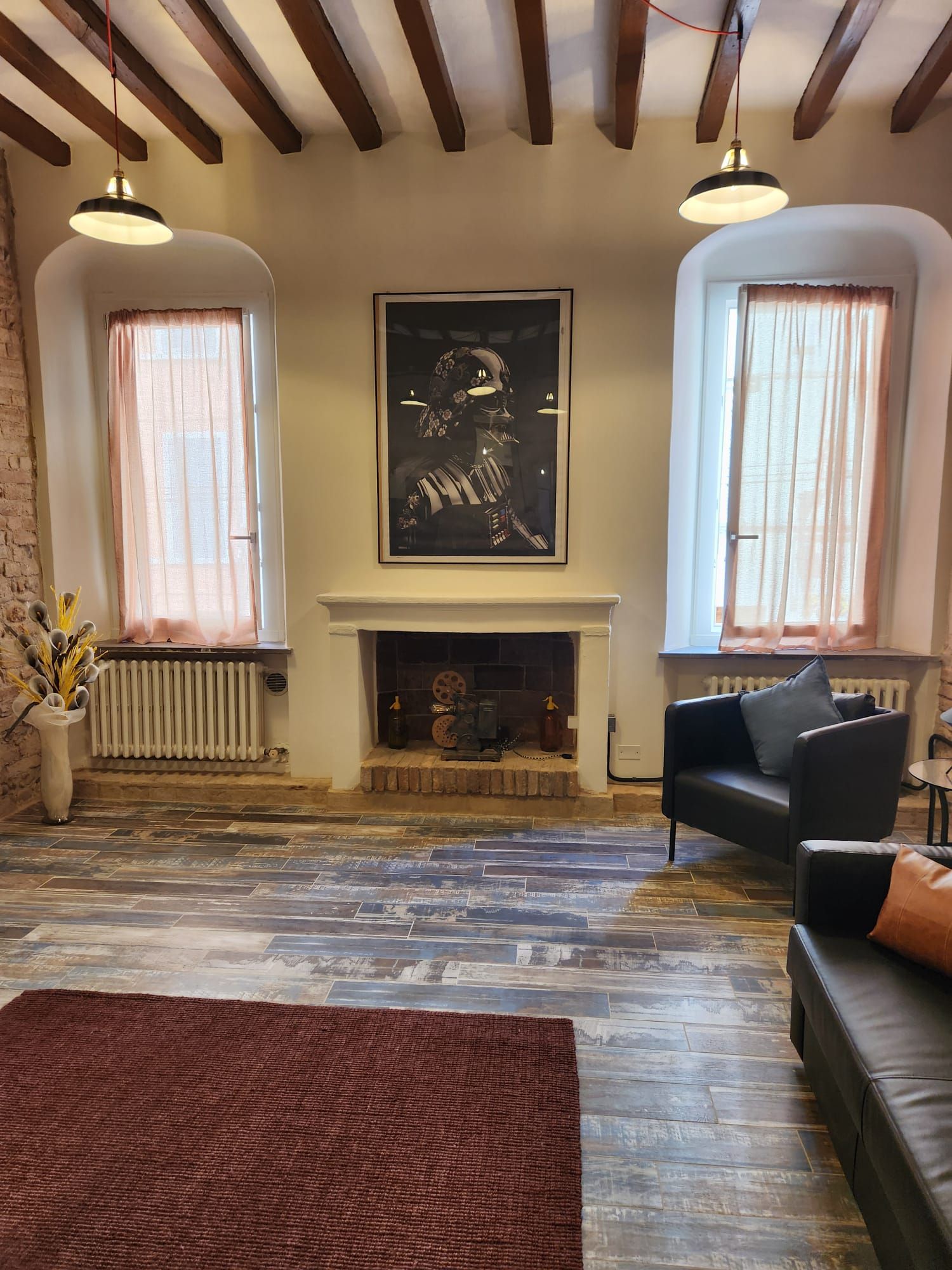 Ein Wohnzimmer mit Kamin, zwei Fenstern mit transparenten Vorhängen, einem schwarzen Sofa und einem braunen Teppich.