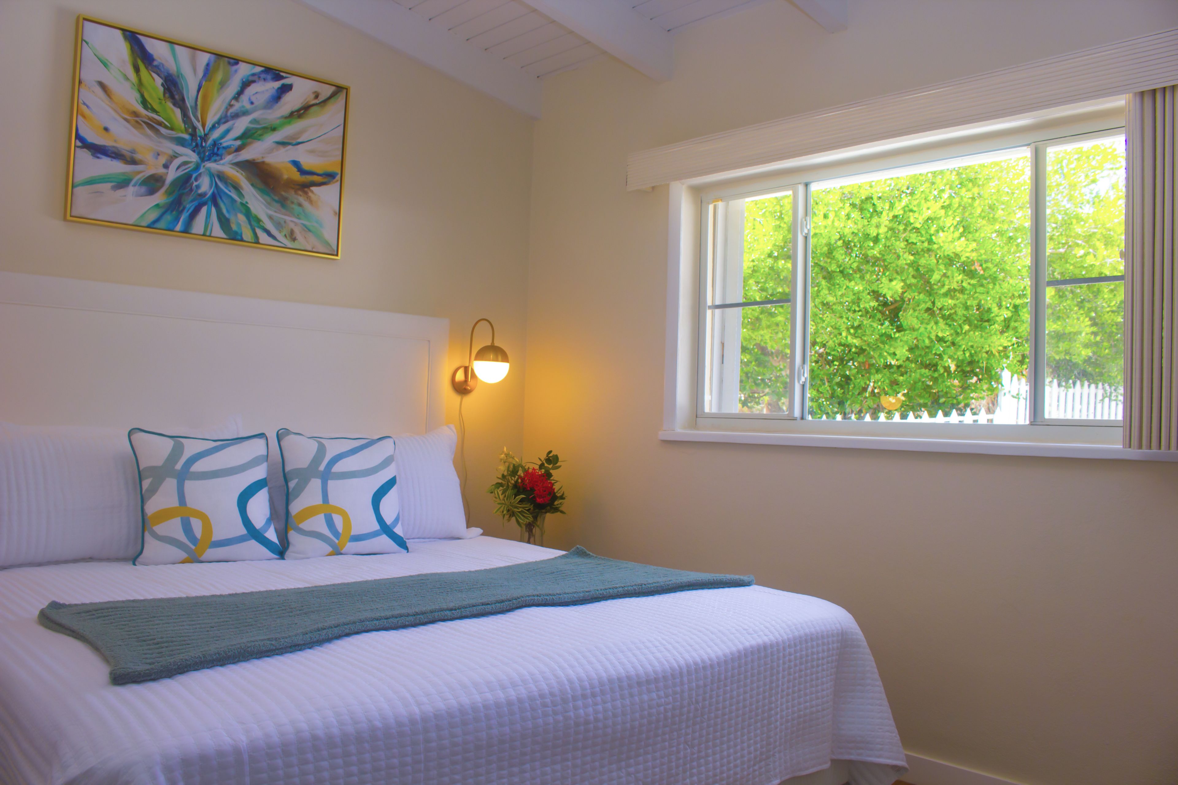 A neatly made bed with a quilt and pillows, a wall-mounted light, a window with a view of trees, and a painting on the wall.