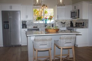 A modern kitchen with white cabinetry, stainless steel appliances, a central island with bar stools, and a view of the outdoors through a window. - 3-bedroom East End Villa with Pool & amazing views of the BVI islands & St John (St Thomas)