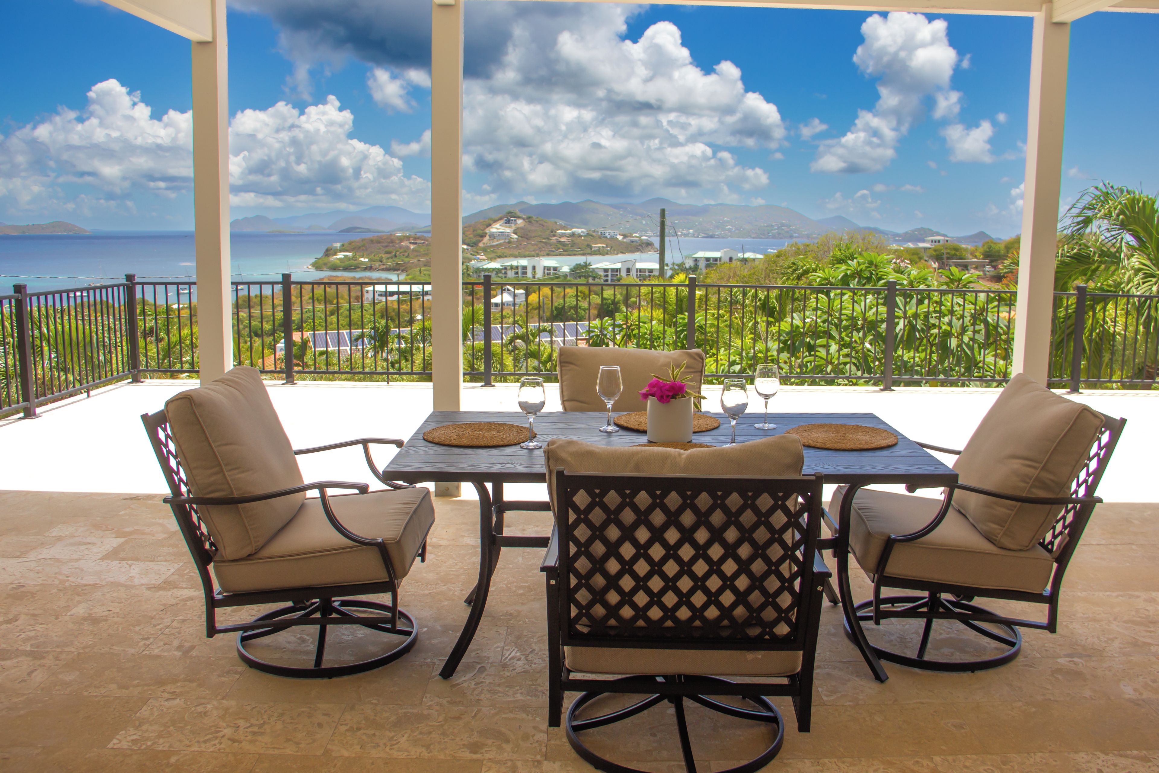 A covered outdoor dining area with a table set for two, surrounded by chairs, overlooking a scenic view of the ocean and hills.