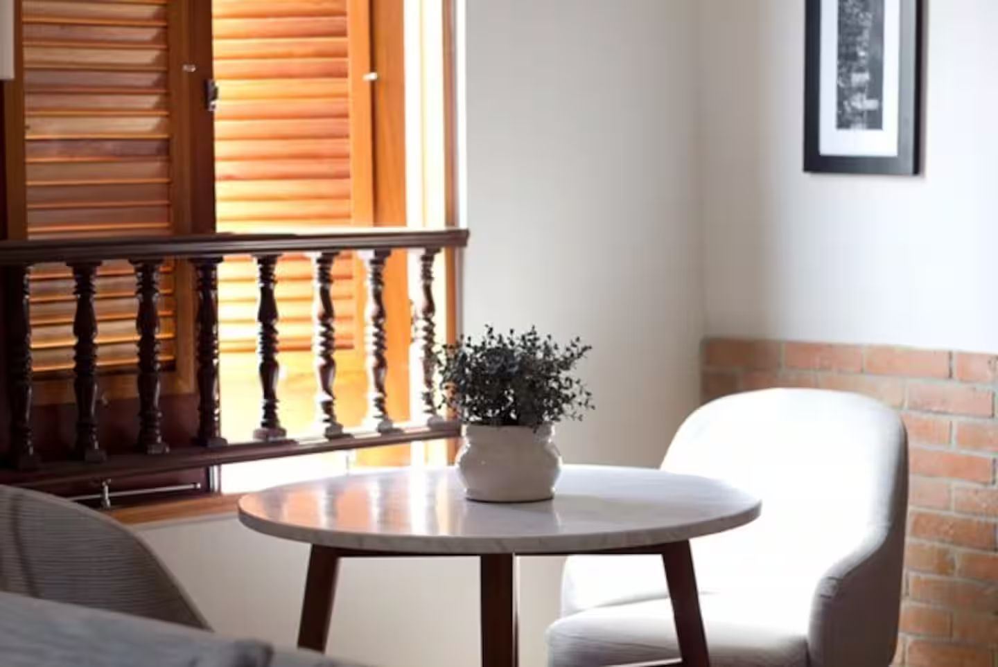 A room with a round table, a potted plant, a white chair, a wooden railing, and a brick wall.