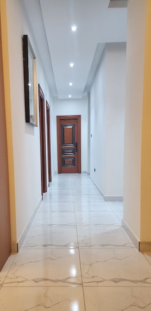 Un pasillo con piso de mármol, una puerta de madera al final y luces empotradas en el techo.