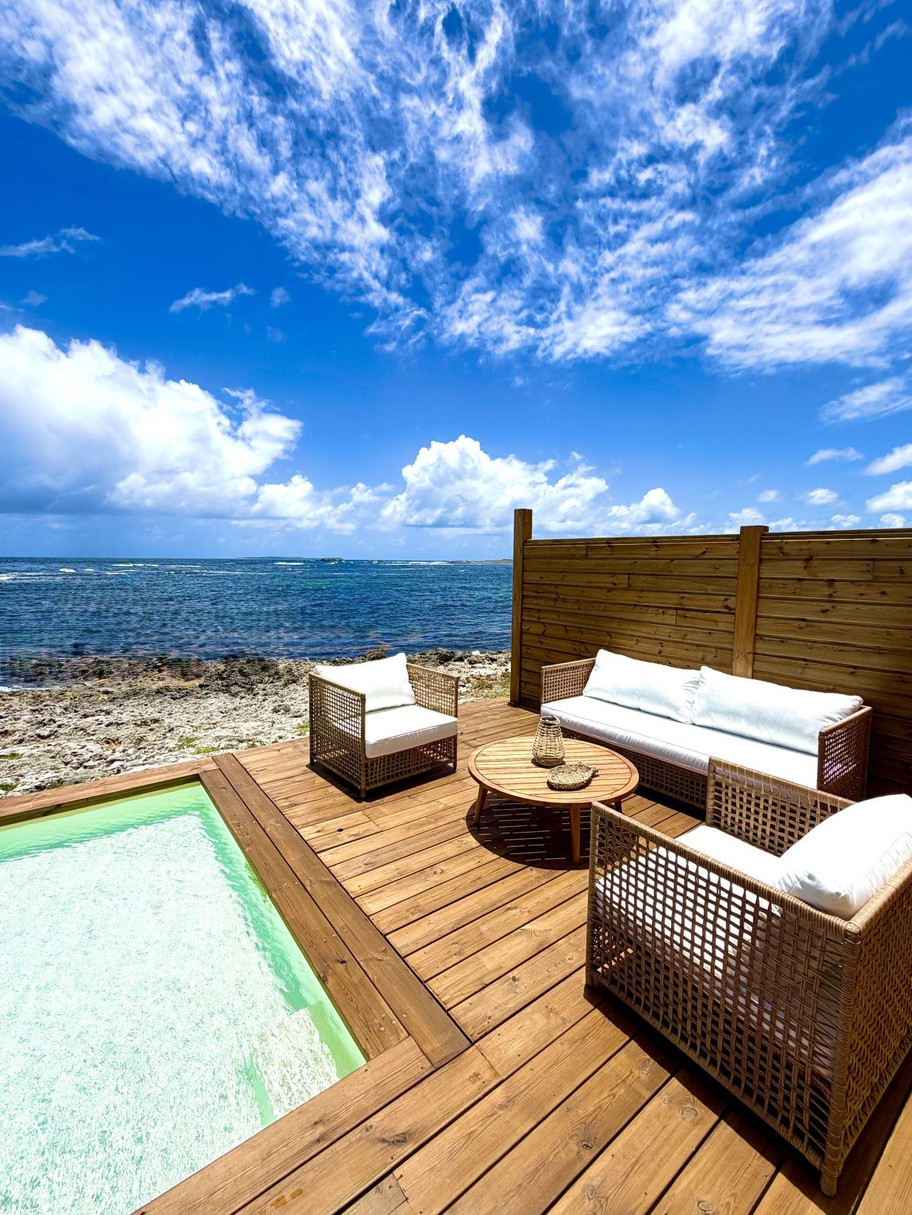 A wooden deck with wicker furniture and a pool, overlooking a beach and ocean.