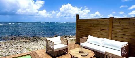 Terraza de madera con mobiliario de mimbre y piscina, con vistas a una playa y al océano.