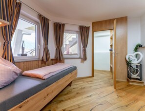 Ein Schlafzimmer mit einem hölzernen Bett, zwei Fenstern mit Vorhängen, einer Holztür und einem kleinen Regal mit einer herzförmigen Dekoration.