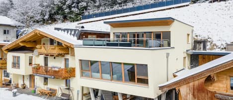 A modern ski resort with chalet-style buildings, a covered outdoor seating area, and a snowy mountain backdrop.