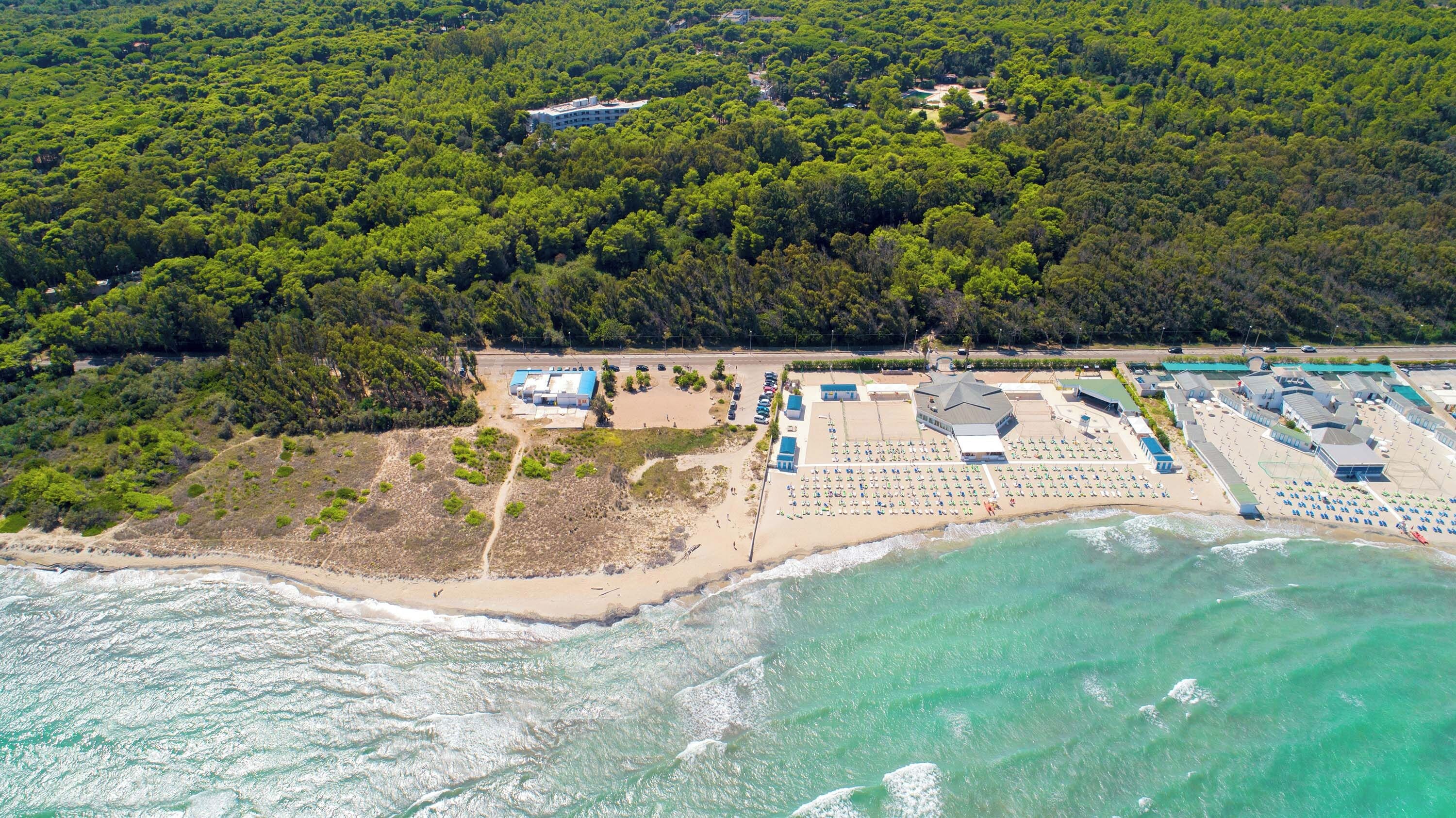 An aerial view of a coastal resort with a beach, a parking area, and a large building with multiple sections.