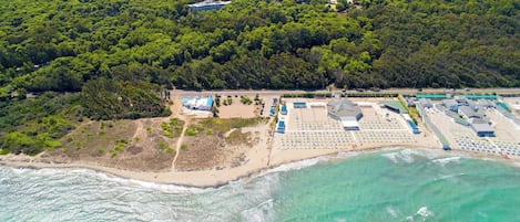 An aerial view of a coastal resort with a beach, a parking area, and a large building with multiple sections.