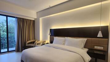 A modern hotel room with a large bed, a ceiling fan, a bedside table, and a view of greenery outside.