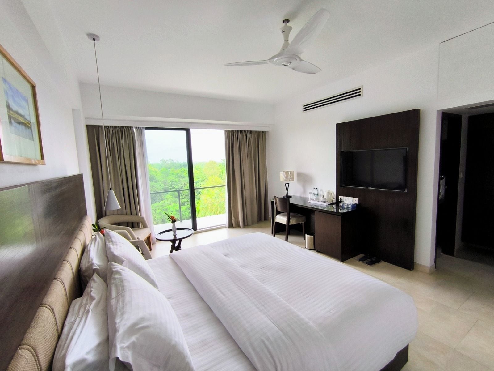 Habitación de hotel con una cama grande, un escritorio con silla, televisor y balcón con cortinas.