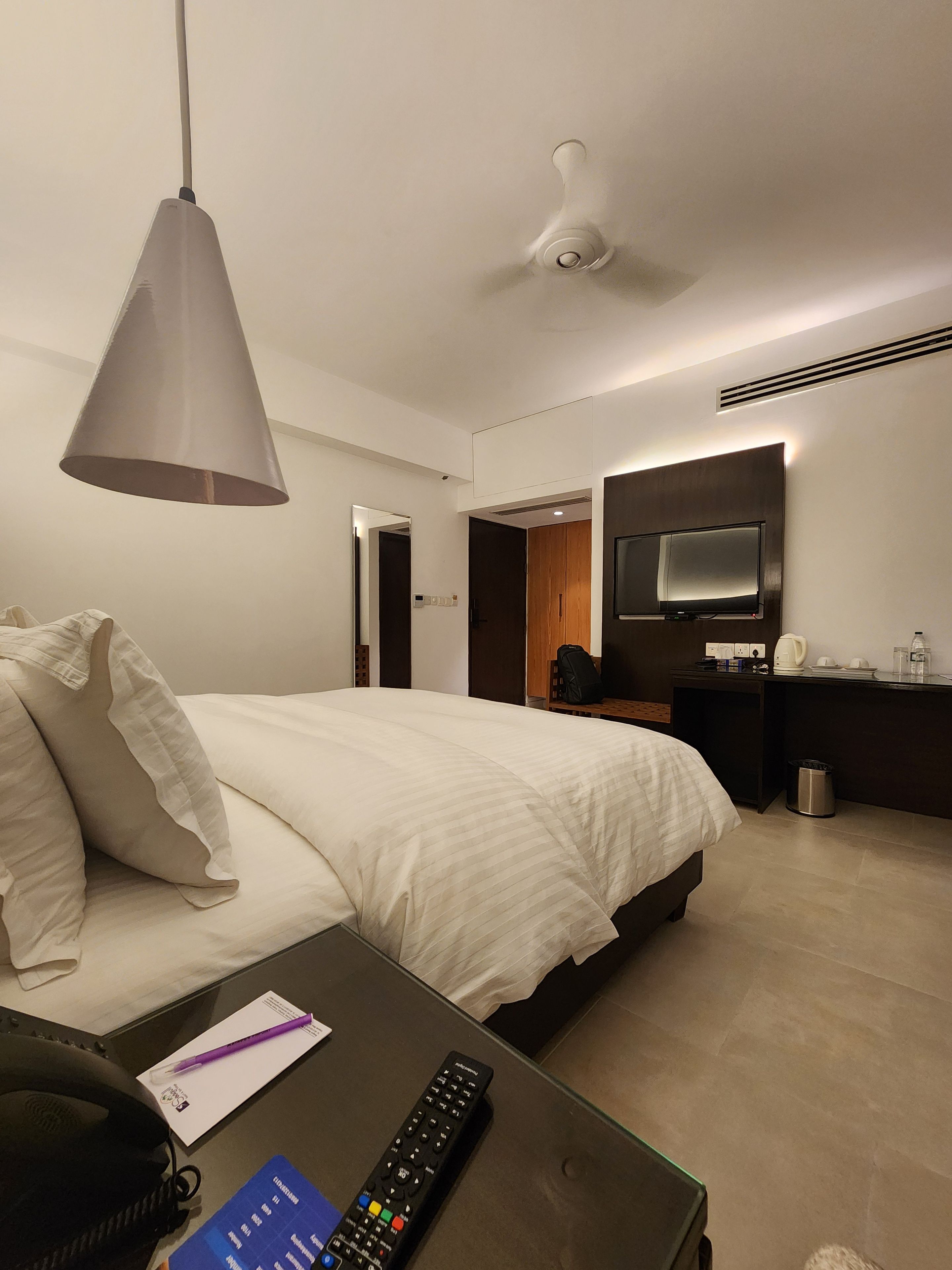 A modern hotel room with a bed, bedside table, lamp, TV, and a small desk with a trash bin.