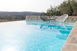 An infinity pool with clear blue water, surrounded by a stone wall and two chairs. - Tranquil 3-bedroom villa with swimming pool and beautiful country views (Sicilia)