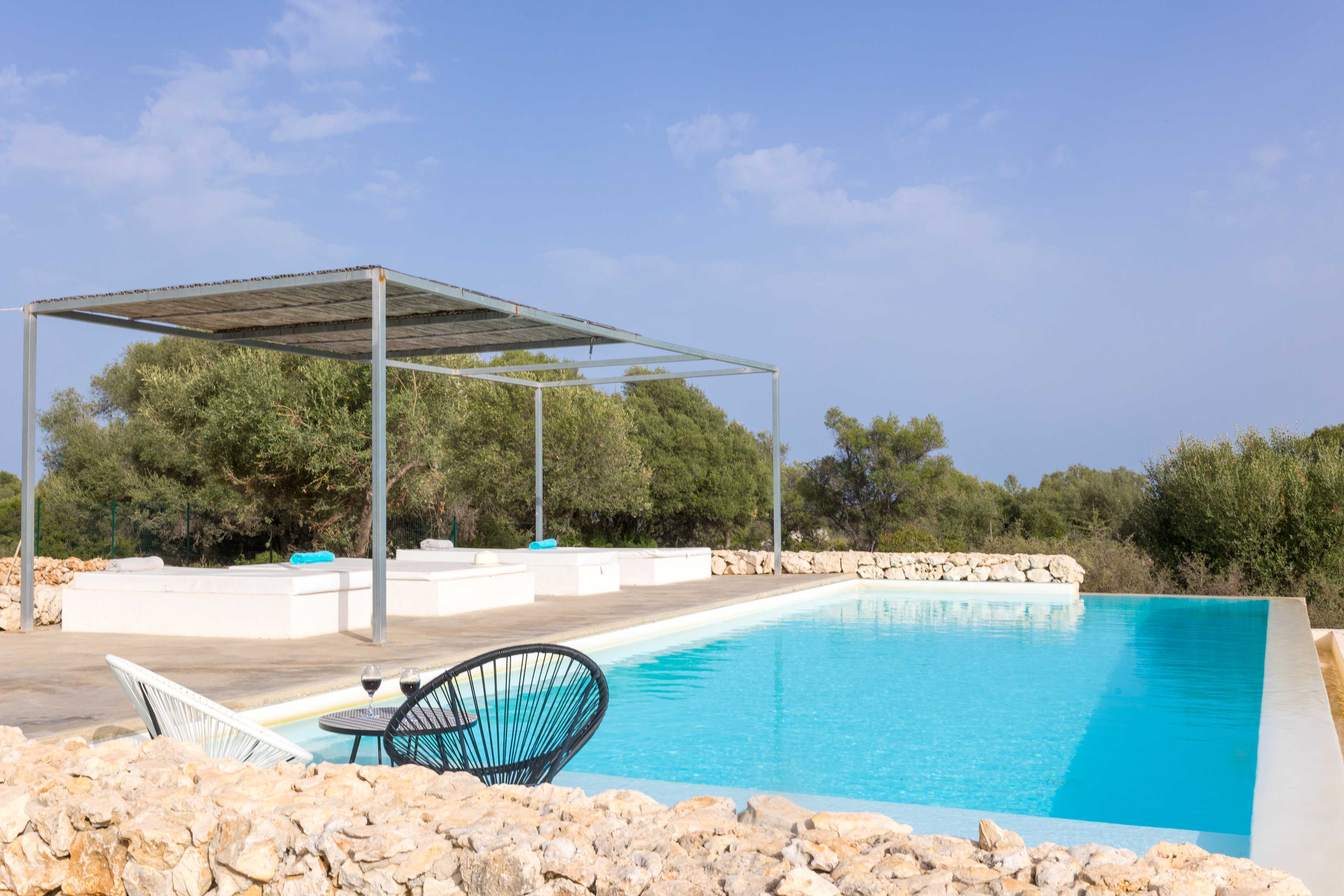 En swimmingpool med en pergola, sittplatser i sten och en stol.