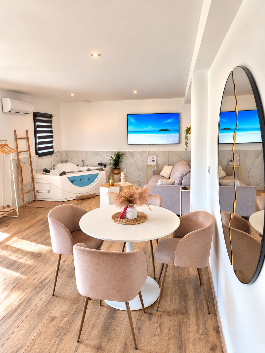 A modern living room with a round table, two chairs, a sofa, a large mirror, and a television displaying a beach scene.