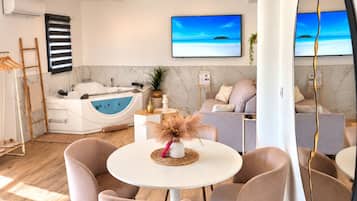 En moderne stue med et rundt bord, to stole, en sofa, et stort spejl og et fjernsyn, der viser et strandmotiv.