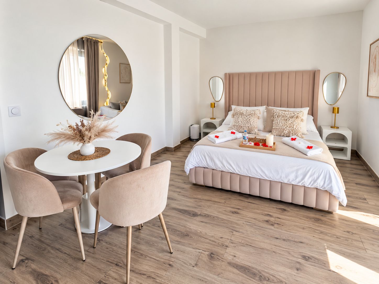 Une chambre moderne avec un grand lit, des tables de chevet, un miroir rond, une table de salle à manger ronde avec des chaises et un vase contenant des fleurs séchées.