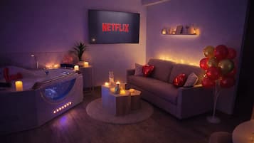 Uma sala de estar aconchegante com uma poltrona de massagem, uma TV de tela plana exibindo 'NETFLIX', um sofá com almofadas em formato de coração e uma mesinha com velas.