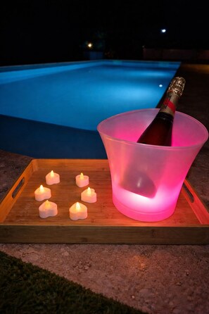 Escena junto a la piscina con una bandeja de madera que sostiene una botella de champán y varias velas encendidas.