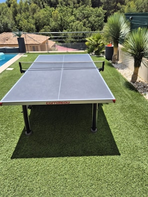 A ping pong table with a net in a backyard with artificial grass, surrounded by trees and a pool.