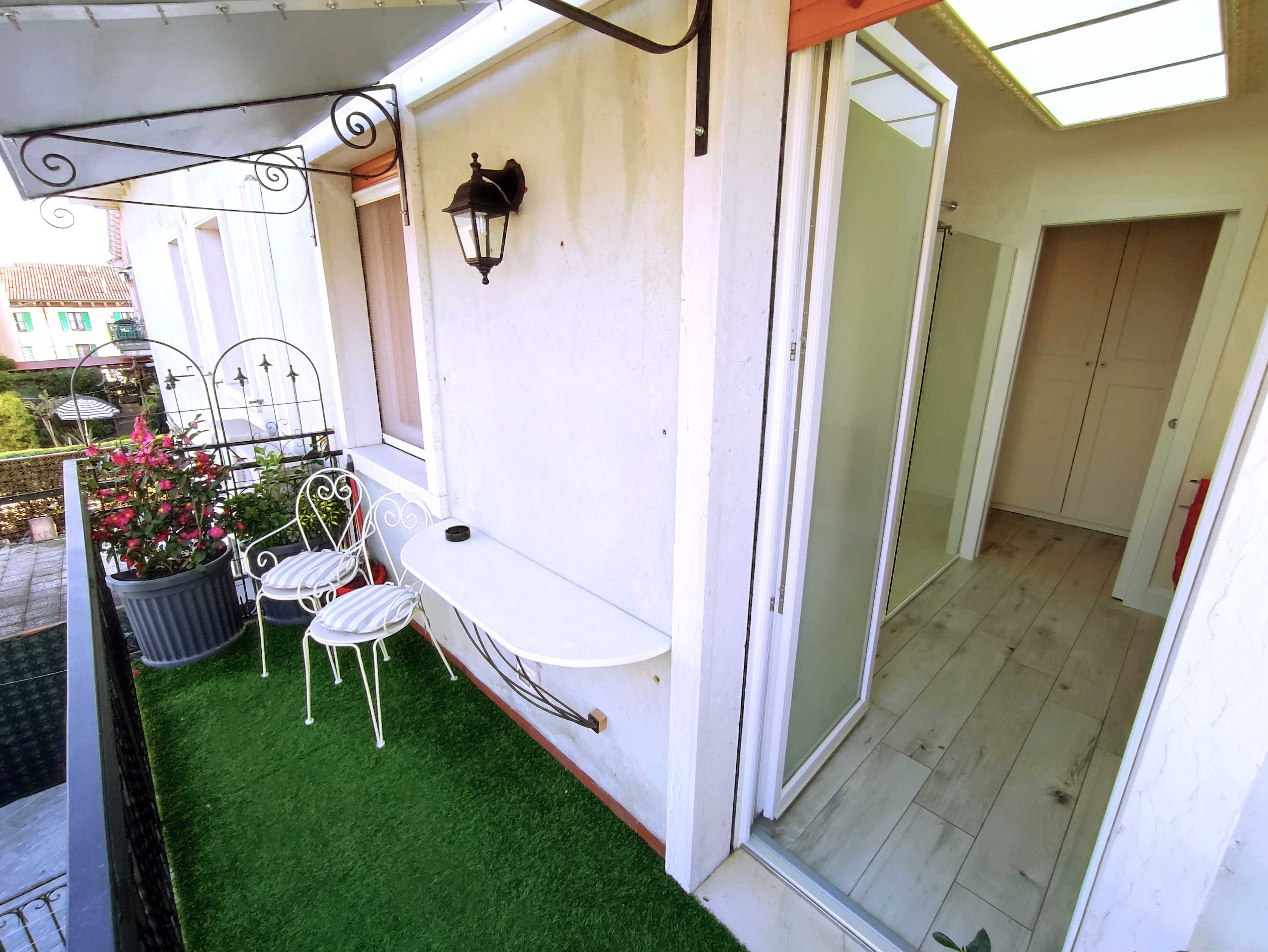 Ein Balkon mit Kunstgrasanlage, einem weißen Tisch, zwei Stühlen, einem Blumentopf mit rosa Blüten und einer Wandleuchte.