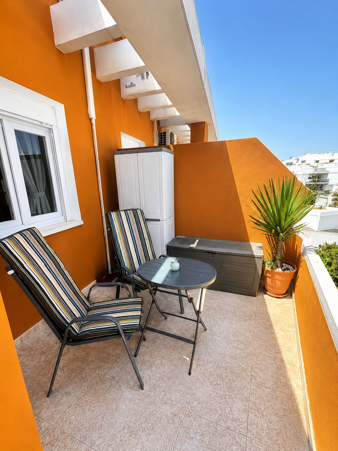 Un balcon avec deux chaises à rayures, une petite table ronde, une plante en pot et un meuble blanc.
