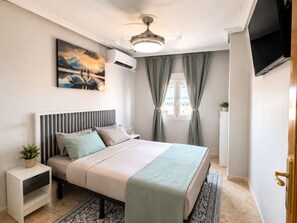 A bedroom with a bed, a nightstand, a ceiling fan, an air conditioning unit, a window with curtains, and a TV mounted on the wall. - Modern Coastal Penthouse w AC & Lg Private Terrace (Torrevieja)