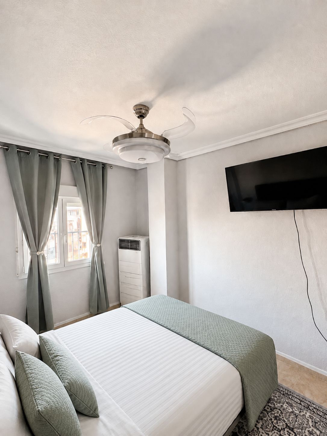 A bedroom with a bed, curtains, a ceiling fan, an air conditioning unit, and a mounted TV.